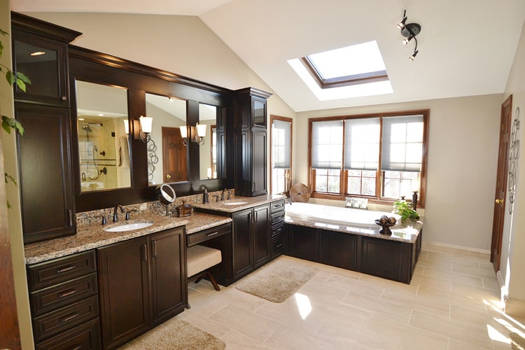 remodeled bathroom with dark cabinets, granite counters, two sinks, a makeup vanity, built-in tub, and skylight