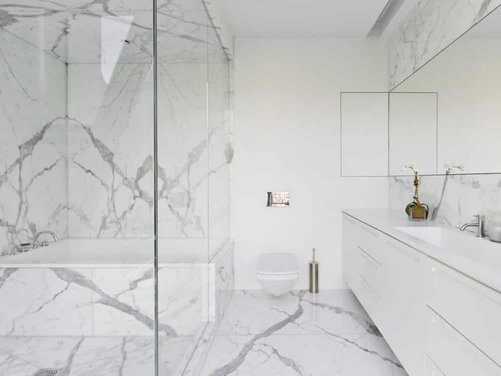 remodeled bathroom featuring marble floors, walls, and countertops with a white vanity and large glass shower with a tub in it