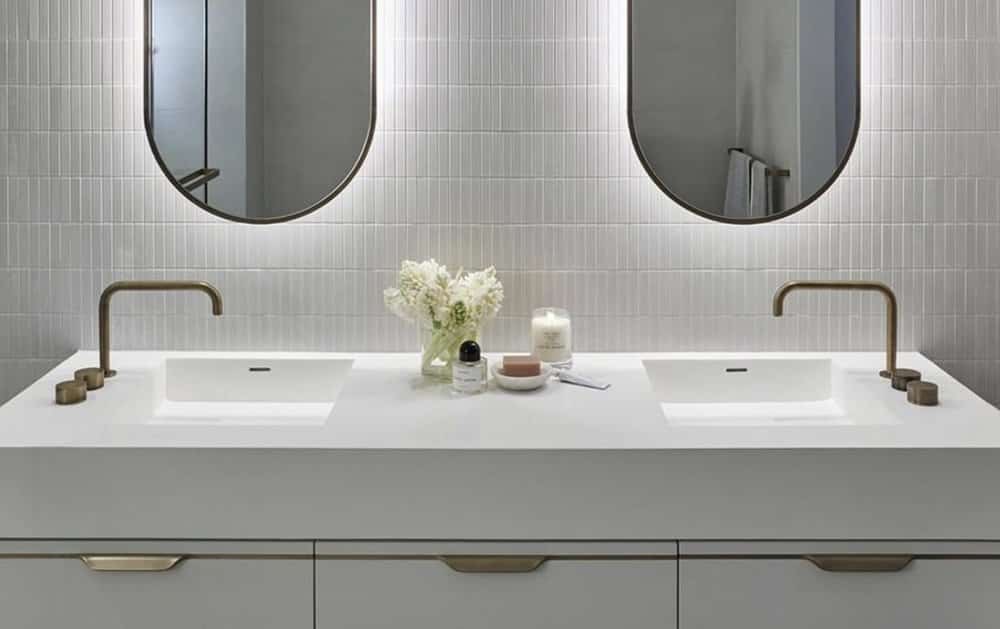 remodeled bathroom with white wall tile, white vanity and countertop, backlit mirrors, and two sinks with side-mounted faucets